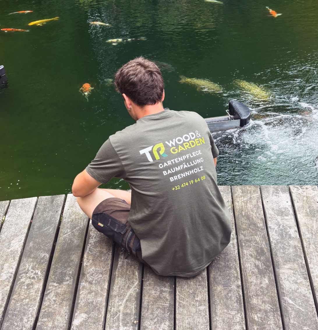 Mann in TR Wood & Garden Shirt sitzt auf Holzsteg und füttert Fische in grünem Wasser.