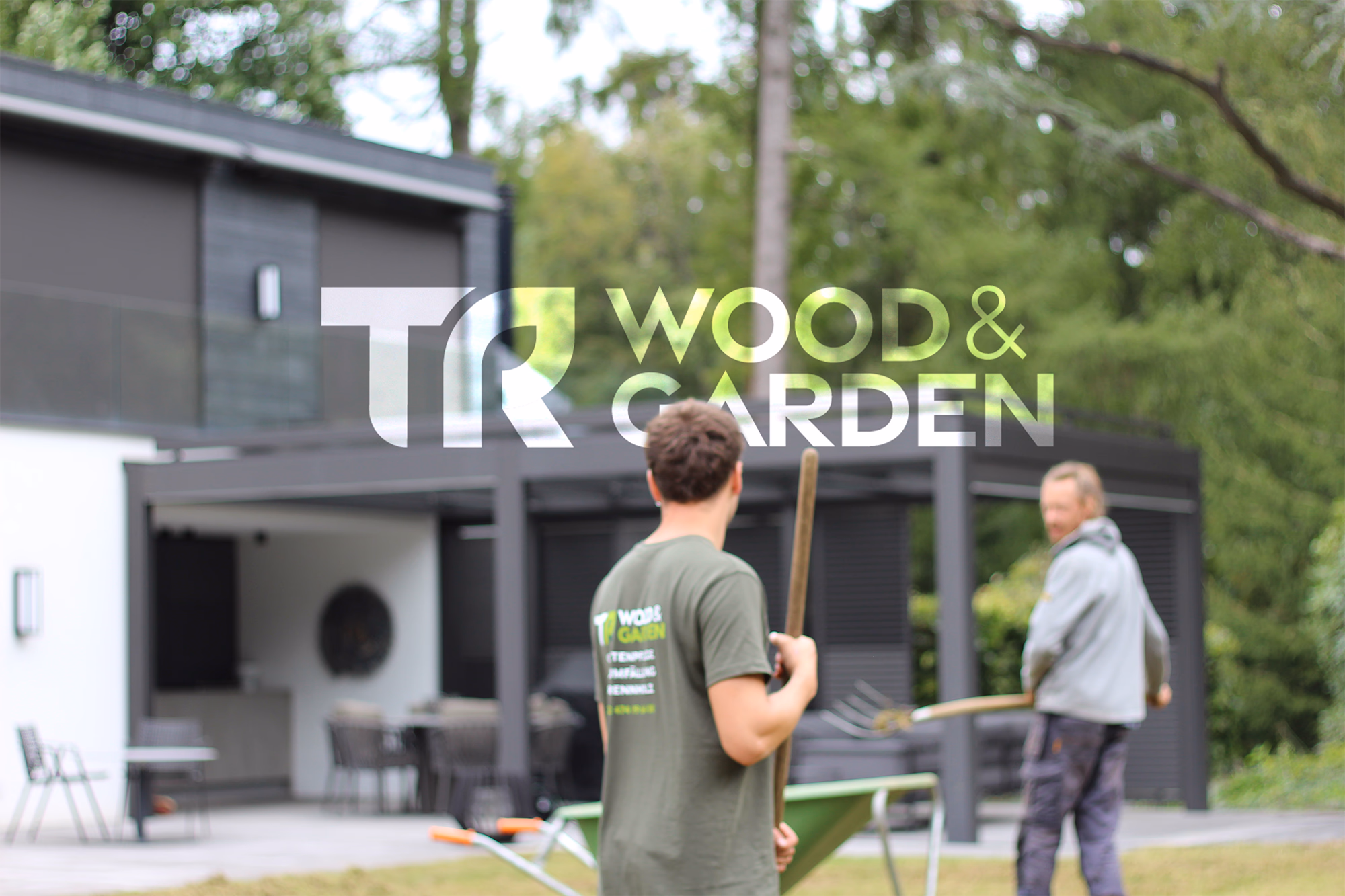 Arbeiter in grünem Shirt mit Schaufel vor modernem Gartenhaus, Kollege im Hintergrund, grüne Bäume sichtbar.