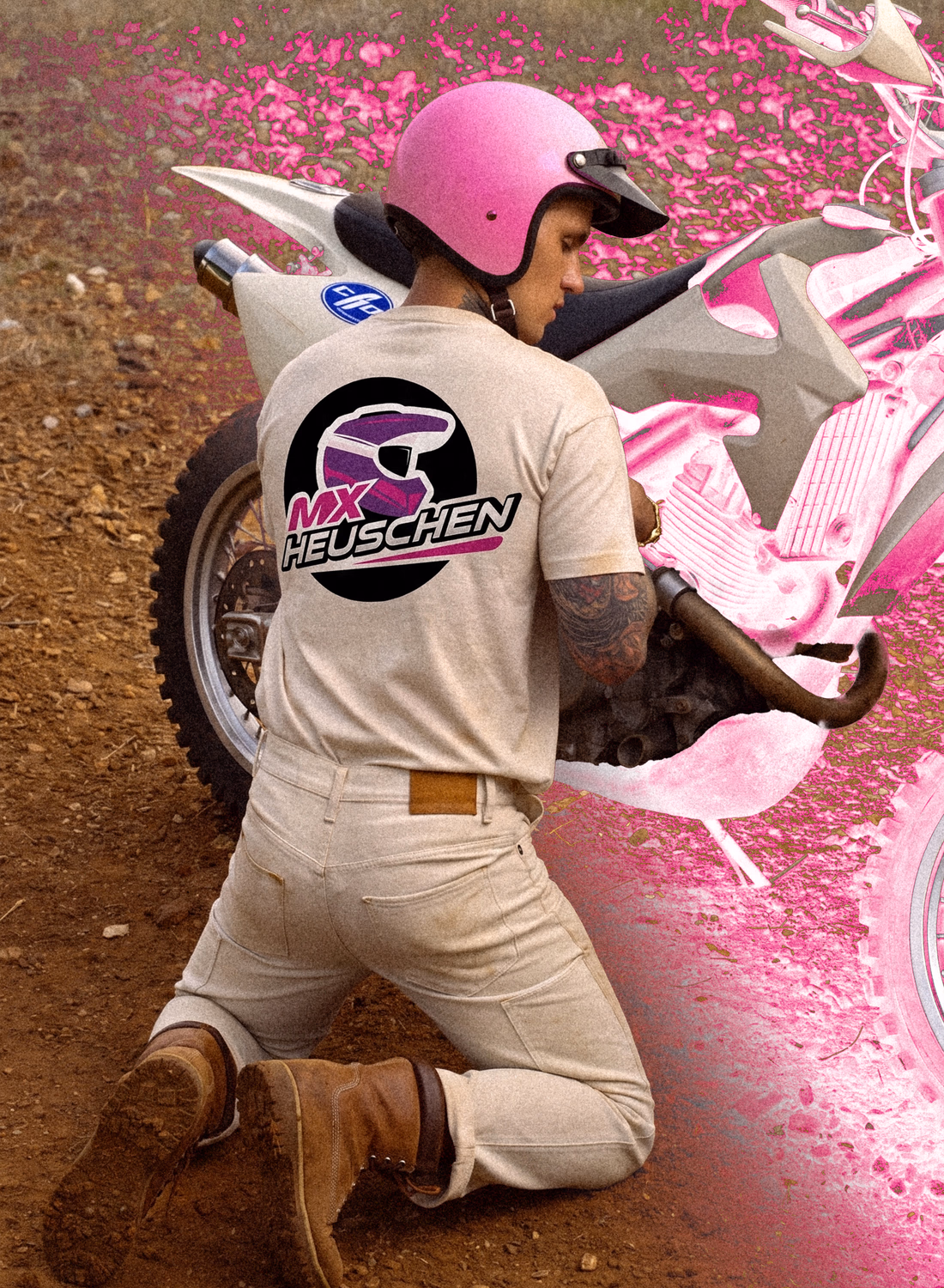 Motorradfahrer in rosa Helm und beigem MX Heuschen Shirt kniet vor pinker Blumenkulisse.